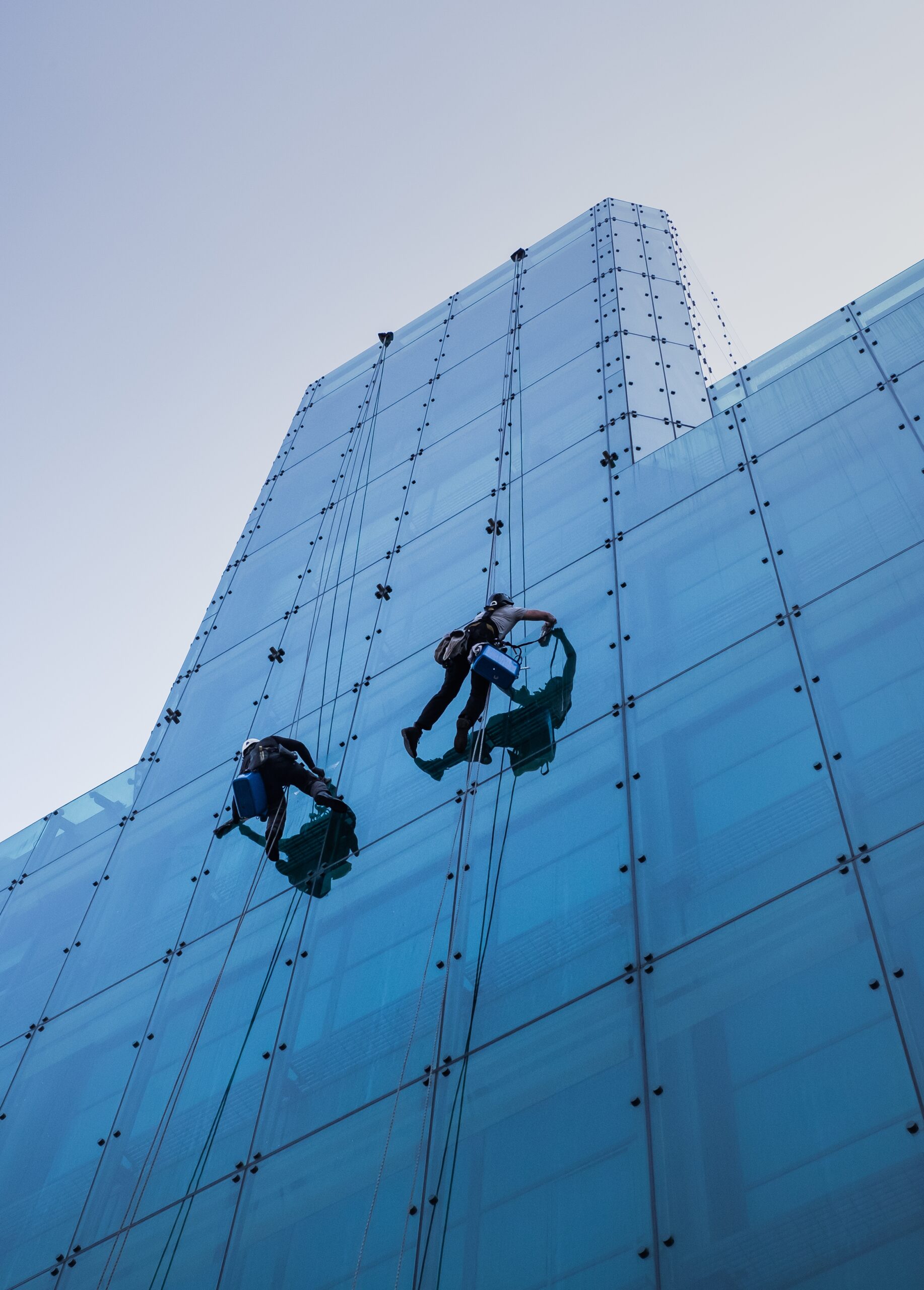 vertical low angle shot of two people climbing a tall glass building during the day time